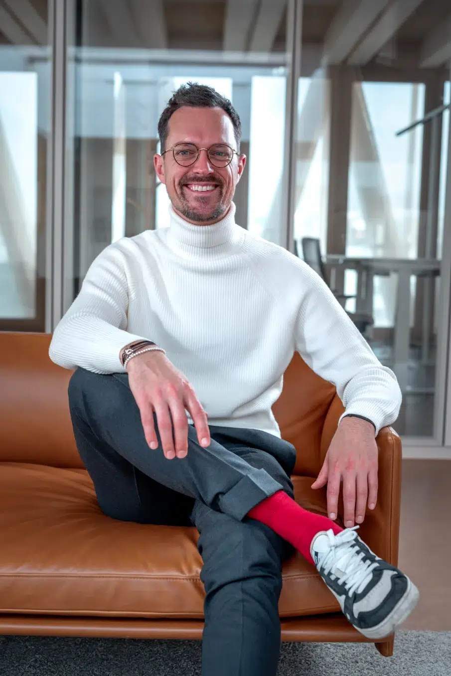 Mann mit Brille und weißem Rollkragenpullover sitzt entspannt auf einem Ledersofa in modernem Büro – Fördermittelberatung Experte.